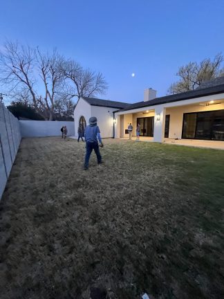 A "before" construction photo showing two contractors surveying a large, dormant grass backyard at dusk. The site features a modern white stucco home with a prominent arched glass door and a light gray horizontal fence, capturing the initial planning phase of a custom pool and landscape transformation.