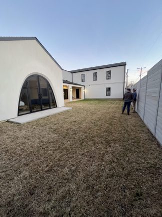 A "before" construction photo showing two contractors surveying a large, dormant grass backyard at dusk. The site features a modern white stucco home with a prominent arched glass door and a light gray horizontal fence, capturing the initial planning phase of a custom pool and landscape transformation.