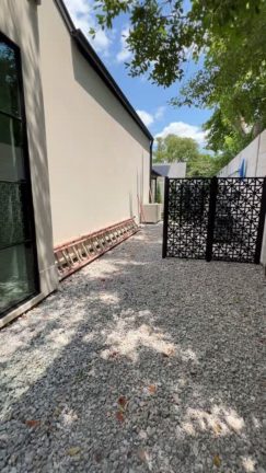 A side yard with a gravel ground cover containing pool equipment and a beige utility box. A black decorative laser-cut metal screen partially hides the pool filter and pipes. In the background, a small lawn area is visible, bordered by a white fence and tall evergreen trees under a blue sky.
