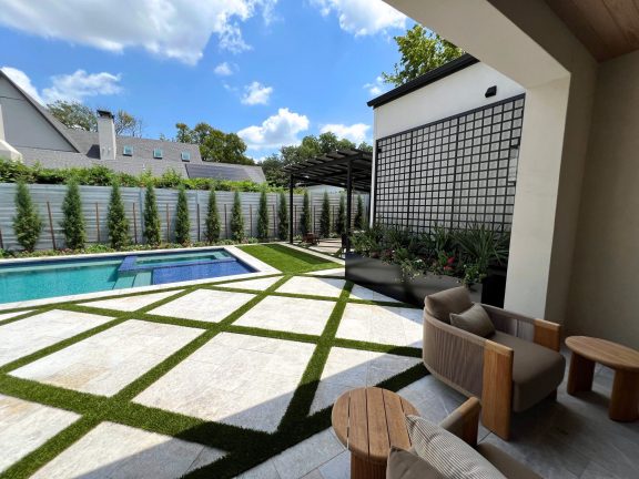 A modern backyard featuring a geometric stone and turf patio beside a rectangular swimming pool. In the shaded foreground, contemporary lounge chairs and wooden side tables sit near a black planter and trellis, while tall cypress trees line the perimeter under a bright, sunny sky.
