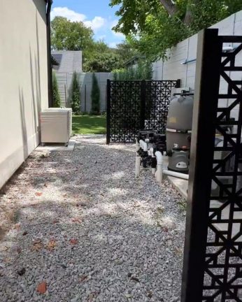 A side yard with a gravel ground cover containing pool equipment and a beige utility box. A black decorative laser-cut metal screen partially hides the pool filter and pipes. In the background, a small lawn area is visible, bordered by a white fence and tall evergreen trees under a blue sky.