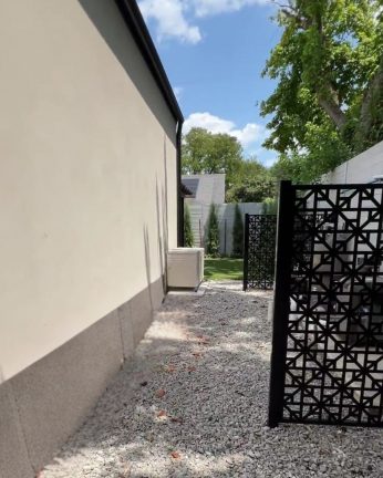 A finished side yard project featuring crushed gray gravel and decorative black laser-cut metal privacy screens. The path leads from the side of a modern white stucco home toward a lush green backyard with privacy evergreens, showcasing clean utility area management for a luxury pool property.