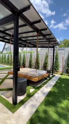 A modern outdoor lounge area featuring a large, wooden hanging daybed suspended by thick ropes from a black metal pergola. The daybed has a white mattress and sits over a geometric patio of light stone pavers and green grass. Tall, slender evergreen trees line a gray fence in the background under a bright, partly cloudy sky.